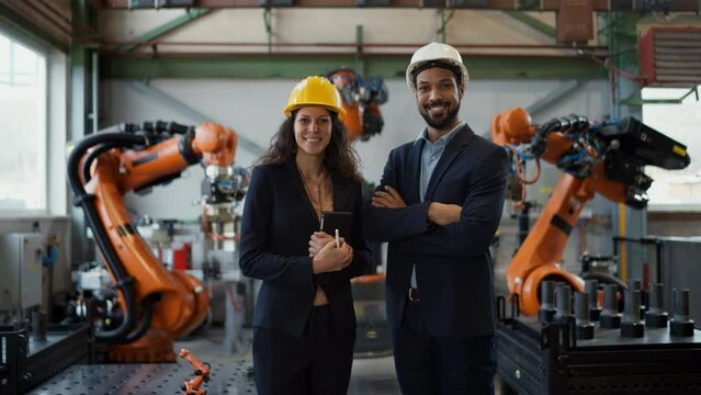 Chief engineer and project manager in modern industrial factory with robot arms looking at camera.