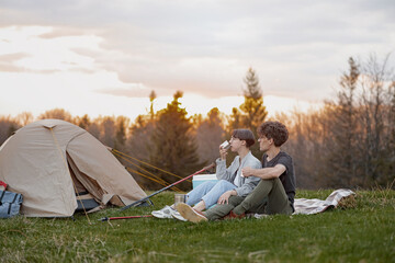 Young couple resting at nature with tent, sitting, talking and drinking tea or coffee.