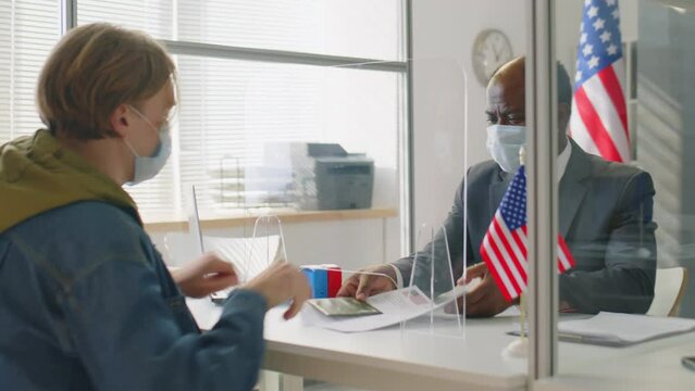 African American consular officer in protective face mask sitting at desk behind glass shield and working visa applicant in US embassy during coronavirus pandemic