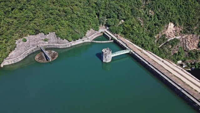 A dynamic aerial shot orbiting the dam and control tower of Shing Mun Reservoir in Hong Kong. There are tourists walking along the pathway across the dam while enjoying the beautiful mountains.