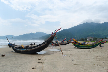 Bent fishing boats on sandy beach Lang Co, Vietnam. Small Vietnamese fishing boats on coast of South China Sea. Green mountains, sea waves and slightly foggy from water splashes in background