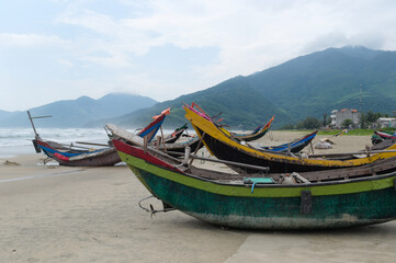 Fototapeta premium Curved fishing boats on coast of South China Sea, Lang Co, Vietnam. Sandy beach with small Vietnamese fishing boats. Green mountains, sea waves and slightly foggy from water splashes in background
