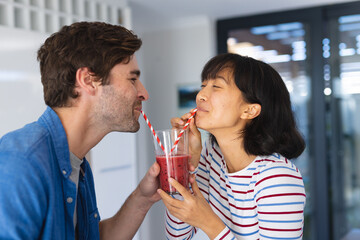 Happy multiracial young couple drinking fruits smoothie through straw in kitchen