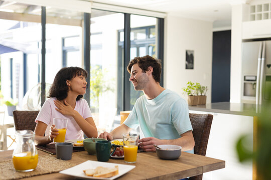 Smiling multiracial young couple talking while having breakfast at dining table in morning