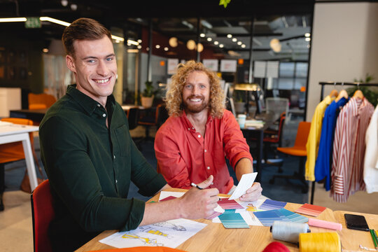 Portrait of smiling caucasian male fashion designers with various fabric swatches at desk in studio