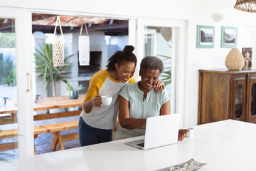 African american young woman with coffee cup standing by mother using laptop on table at home