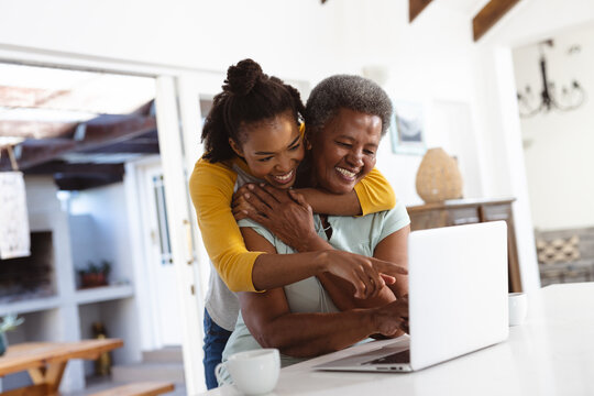 Cheerful african american young woman embracing senior mother using laptop on table at home