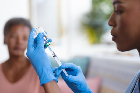 African american senior woman looking at young female doctor holding vial and syringe at home