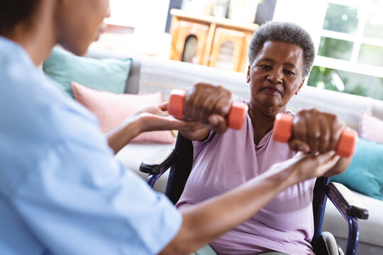 African american doctor assisting senior woman sitting on wheelchair in lifting dumbbells at home