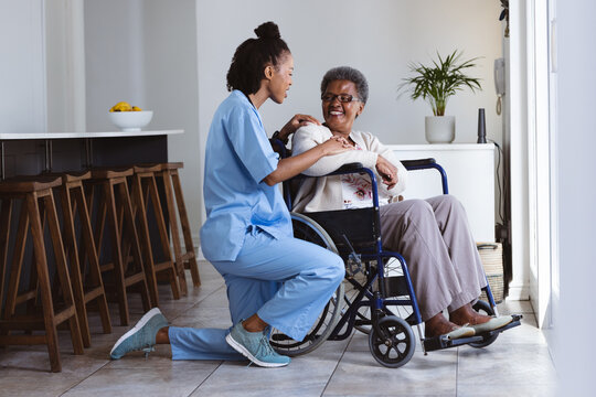 African american female doctor talking with smiling senior patient sitting on wheelchair at home