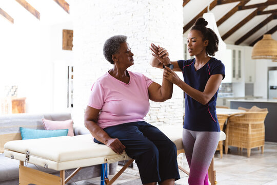 African american female physiotherapist stretching senior woman's wrist sitting on table at home