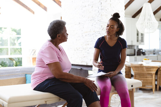 African american female physiotherapist holding clipboard discussing with senior woman at home