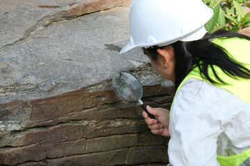 Asian female geologist researcher analyzing rocks with a magnifying glass in a natural park. Exploration Geologist in the Field. Stone and ecology concept. - obrazy, fototapety, plakaty