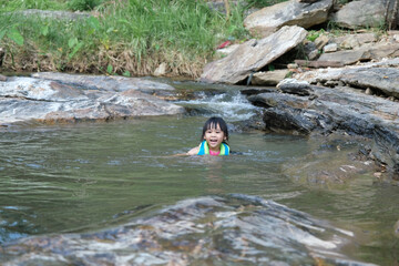 Fototapeta premium Little girls enjoy swimming in the river. Cute Asian girl wearing a life jacket is having fun playing in the stream. Healthy Summer Activities for Kids.