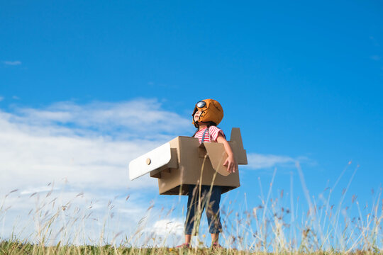 Cute dreamer little girl playing with cardboard planes in the meadow on a sunny day. Happy kid playing with cardboard plane against blue summer sky background. Childhood dream imagination concept.
