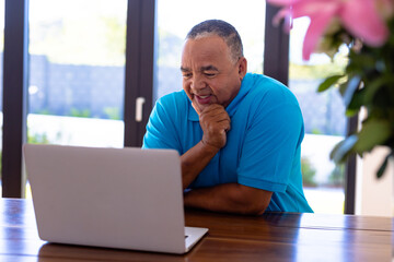 Fototapeta premium Smiling biracial senior man with hand on chin watching video over laptop while sitting at table