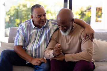 Fototapeta premium Multiracial senior man consoling male friend crying while sitting on sofa in nursing home