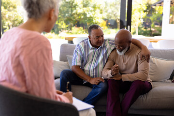 Multiracial female therapist looking at senior man consoling male friend crying in retirement home