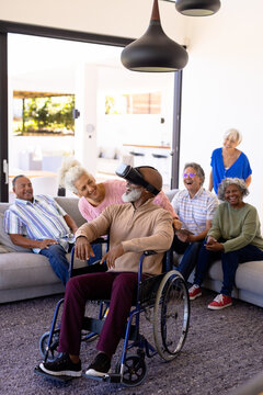Cheerful multiracial seniors looking at friend using virtual reality while sitting on wheelchair