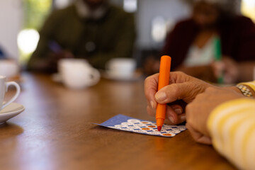 Cropped hands of biracial senior woman marking off numbers on bingo card over table in nursing home