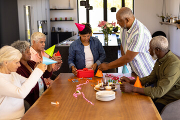 Multiracial seniors wearing party hats while woman opening birthday gift at table in nursing home