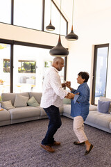 Side view of happy biracial senior friends doing salsa dance by sofa in nursing home, copy space