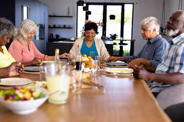 Fototapeta na wymiar Multiracial senior friends with food on dining table holding hands and saying grace in nursing home
