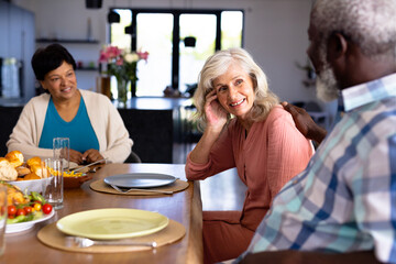 Happy multiracial senior friends with food on dining table talking while sitting in nursing home