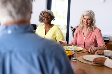 Happy multiracial senior friends talking while sitting at dining table in nursing home