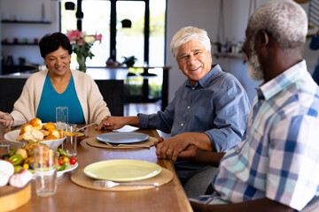 Multiracial smiling senior friends holding hands and talking at dining table in nursing home