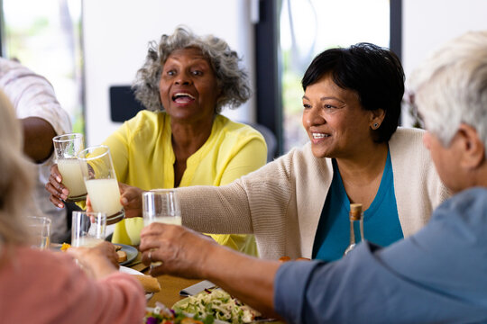 Multiracial cheerful senior friends toasting drinks while having lunch at dining table