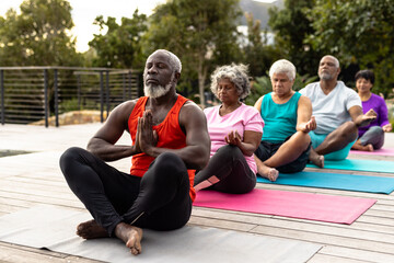 Multiracial senior friends meditating while sitting on mats against plants in yard at nursing home