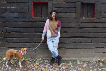 Mujer de pie delante de una cabaña de madera con su perro. Chica con gorro mirando a cámara...