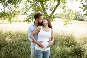 pretty young pregnant woman with white shirt stands with her boyfriend with beard and blue shirt in high flower meadow and cuddle