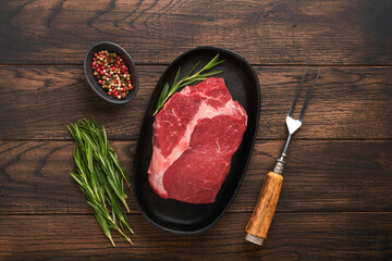Beef steak. Fresh raw beef rib eye steak with fork rosemary, salt and pepper on a grill pan on dark old wooden table background. Shop advertising idea. Top view. Mockup for design idea.