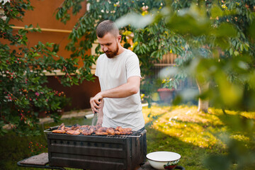A young man in a white t-shirt with a beard makes a barbecue on the grill in the garden near his...