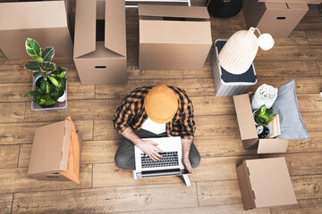 Hipster man with moving boxes in new modern apartment. Happy mature man sitting on the floor of a newly rented ore purchased apartment using a laptop. Unpacked cardboard boxes and furniture in a