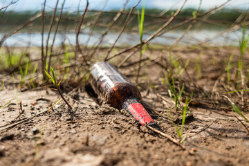 Fototapeta premium Abandoned garbage in nature. Environmental damage caused by garbage pollution. Left glass bottle among the grass against the background of the river