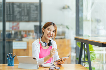 Attractive office worker writing in notepad placed on bright desktop, Female student taking notes from a book at library. Young asian woman sitting at table doing assignments in college library.