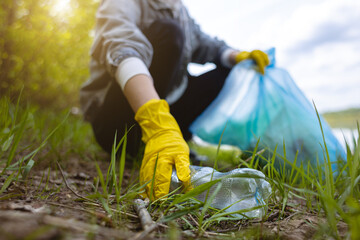 A young woman in yellow gloves collects abandoned garbage in a black bag in the forest. Plastic pollution and environmental protection