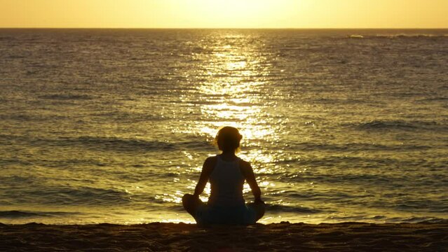 Beautiful silhouette of free woman sitting in meditation pose on beach at golden sunset. Lotus position, medicine yoga asana balance kundalini energy, practice good for woman health and mindfulness