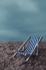 Fototapeta na wymiar A blue and white striped deck chair on sand. Cold moody atmosphere and cloudy sky in background