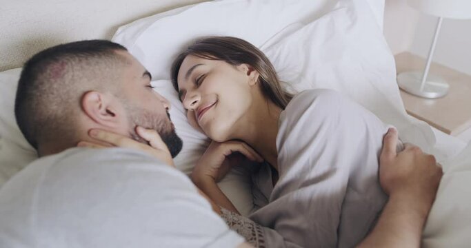 Loving couple lying in bed kissing each other good morning. Happy husband and wife cuddling in bed together in their modern bedroom. Young married couple waking up together in their new home