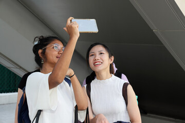Two young attractive Asian multi ethnic woman friends colleagues students talk walk discuss mingle outdoors backpack handphone selfie