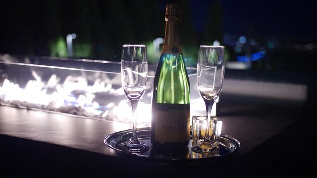 Bottle of Veuve Clicquot champagne on silver tray with two glasses in front of fire at night.
