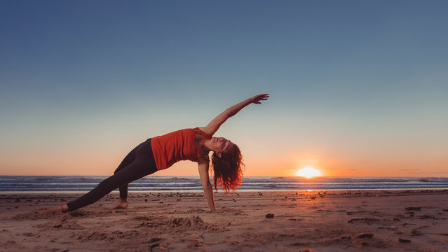 middle-aged woman in sunrise yoga pose in front of the beach with the sun rising from the horizon