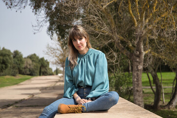 Young, pretty, blonde woman in green shirt, jeans and brown boots, sitting with serious gesture in a park. Concept depression, problems, seriousness, concern.