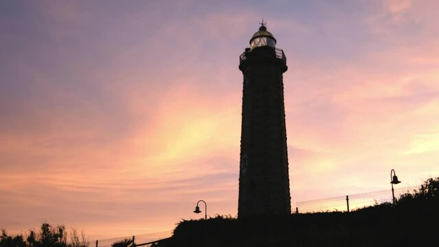 Old Lighthouse guiding with its light the boats that sail in the sea at sunset, with a beautiful landscape of orange clouds. 4K video.