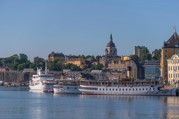New and old steam boats at the pier Skeppsbron in the district Gamla Stan and the church Katarina with old yellow 1700s houses a summer day in Stockholm