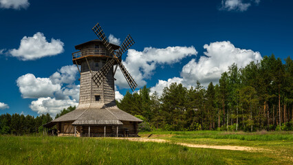 an old wooden windmill on a green meadow in front of a forest under a blue sky with white clouds in fine summer weather